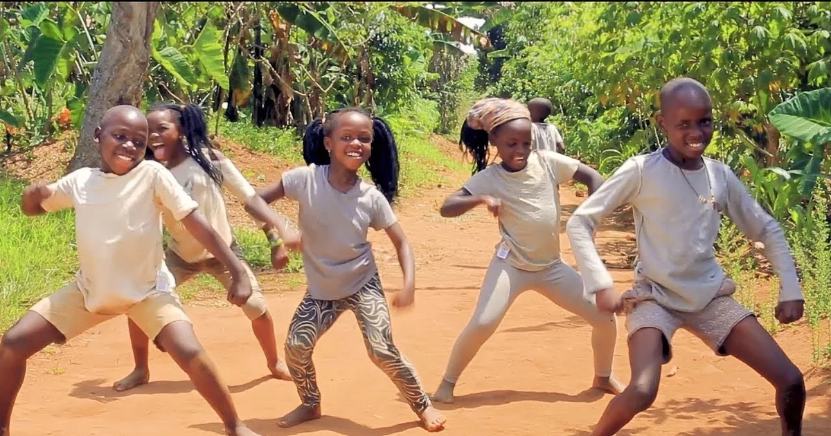 Five people in white shirts and shorts are dancing on a dirt path in a lush green environment.