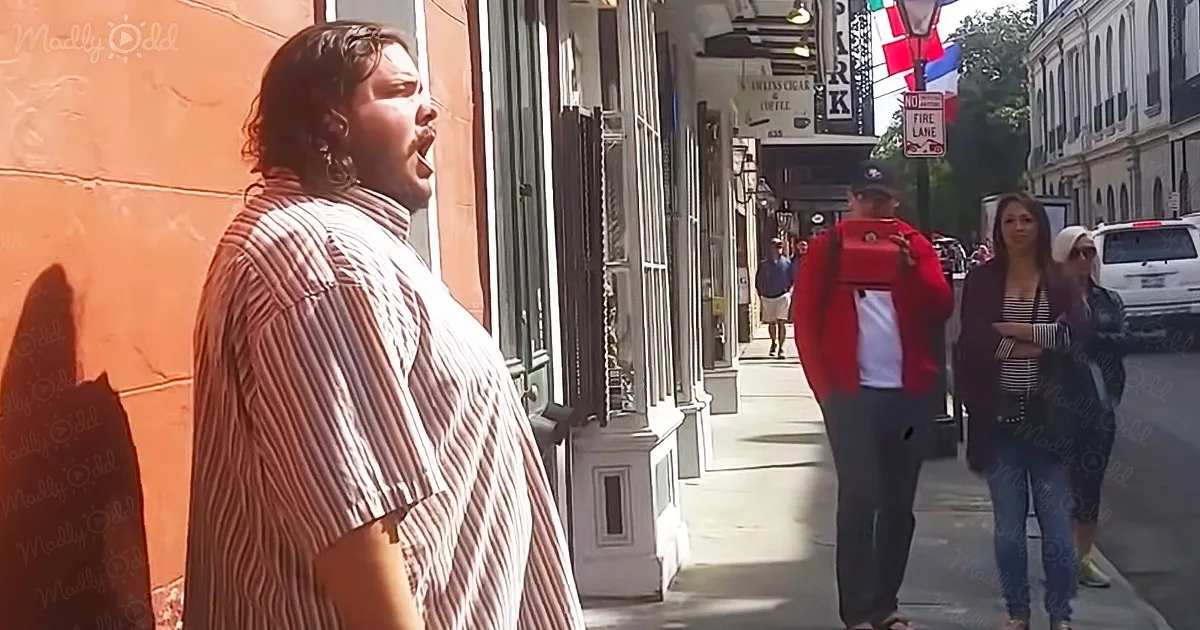 A man in a striped shirt yells on a sunny sidewalk with people and shops in the background.