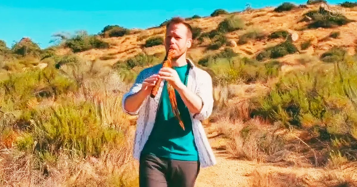 A man in a green shirt and white jacket plays a flute in a grassy, hilly landscape under a clear blue sky.