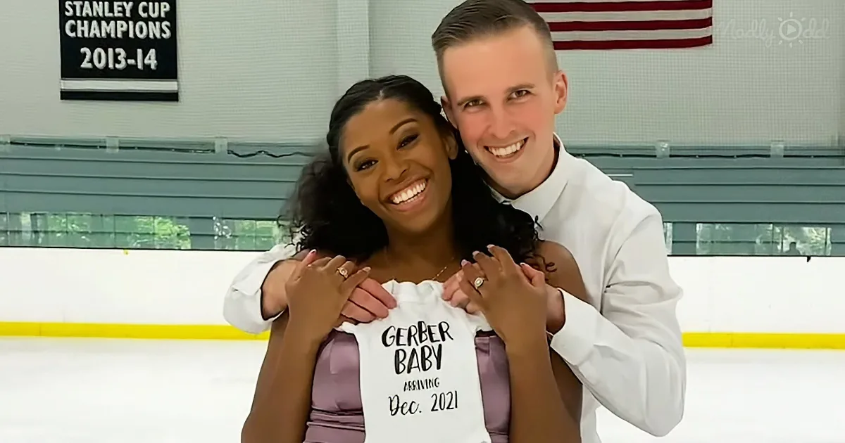 A couple holds a baby onesie announcing their December 2021 arrival at an ice rink.
