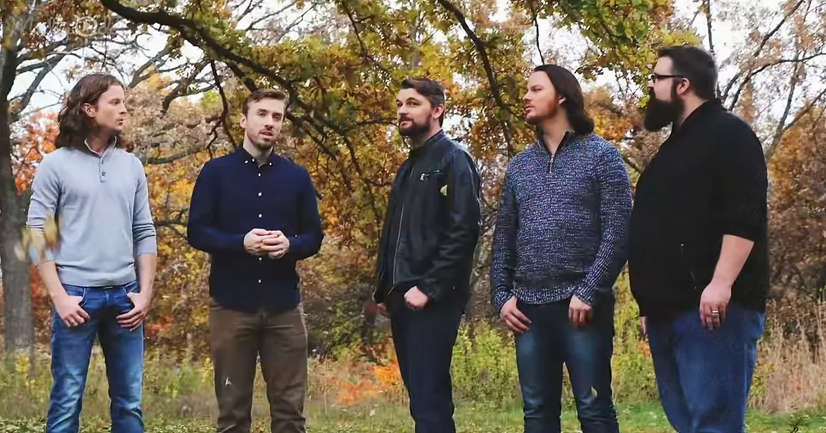 Five men stand outdoors in autumn foliage, dressed casually, seemingly engaged in a discussion.