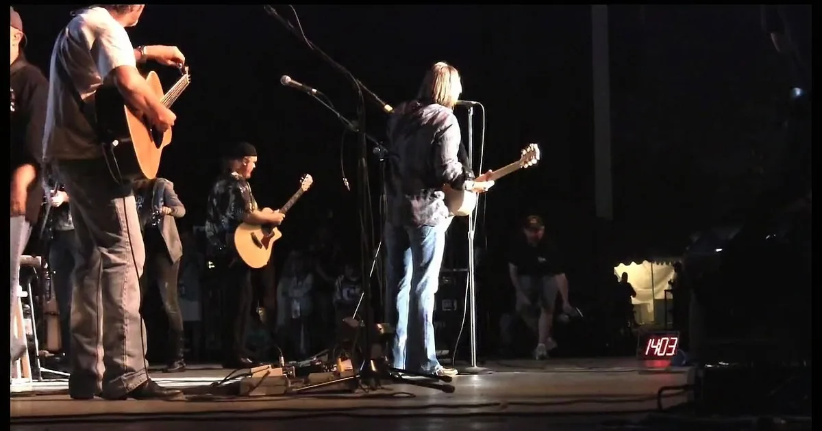 A person in a plaid shirt sings into a microphone while playing guitar on stage.