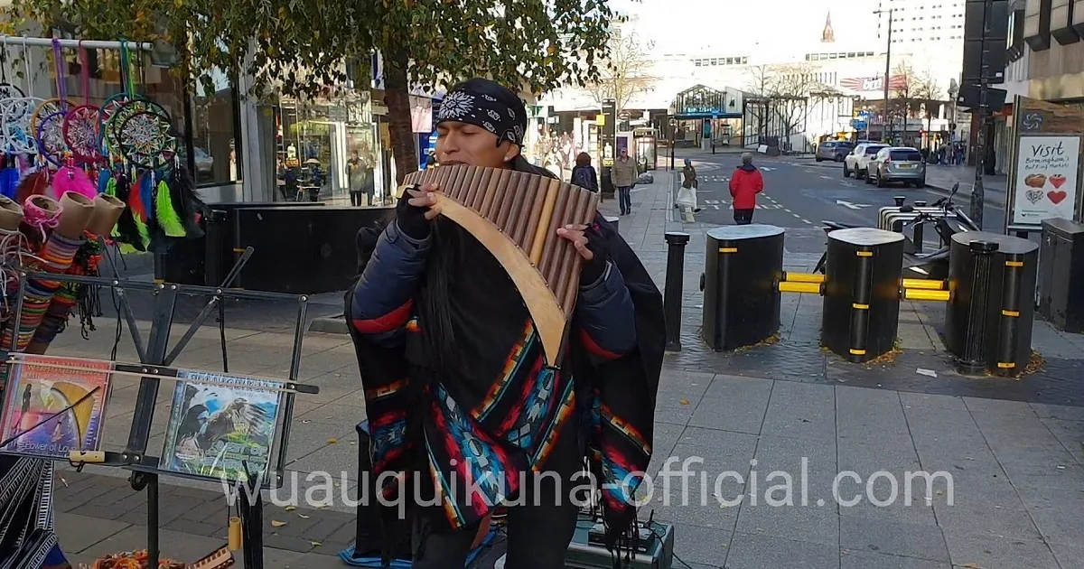 A person plays a long wooden flute against a backdrop of green hills and a cloudy sky.