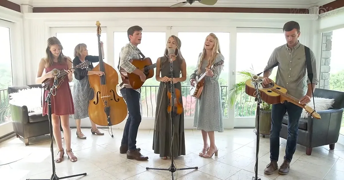A group of musicians perform together, playing various instruments and singing in a bright, open room.