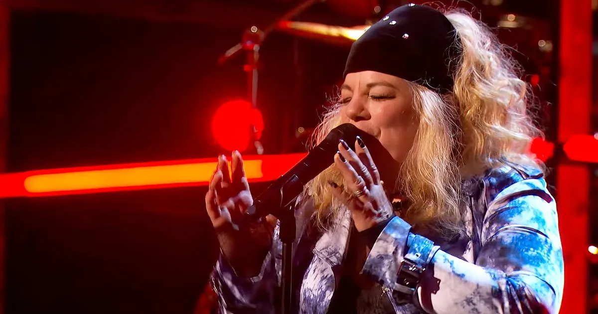 A person with long blonde hair, wearing a black beanie and a patterned jacket, sings passionately into a microphone on stage.