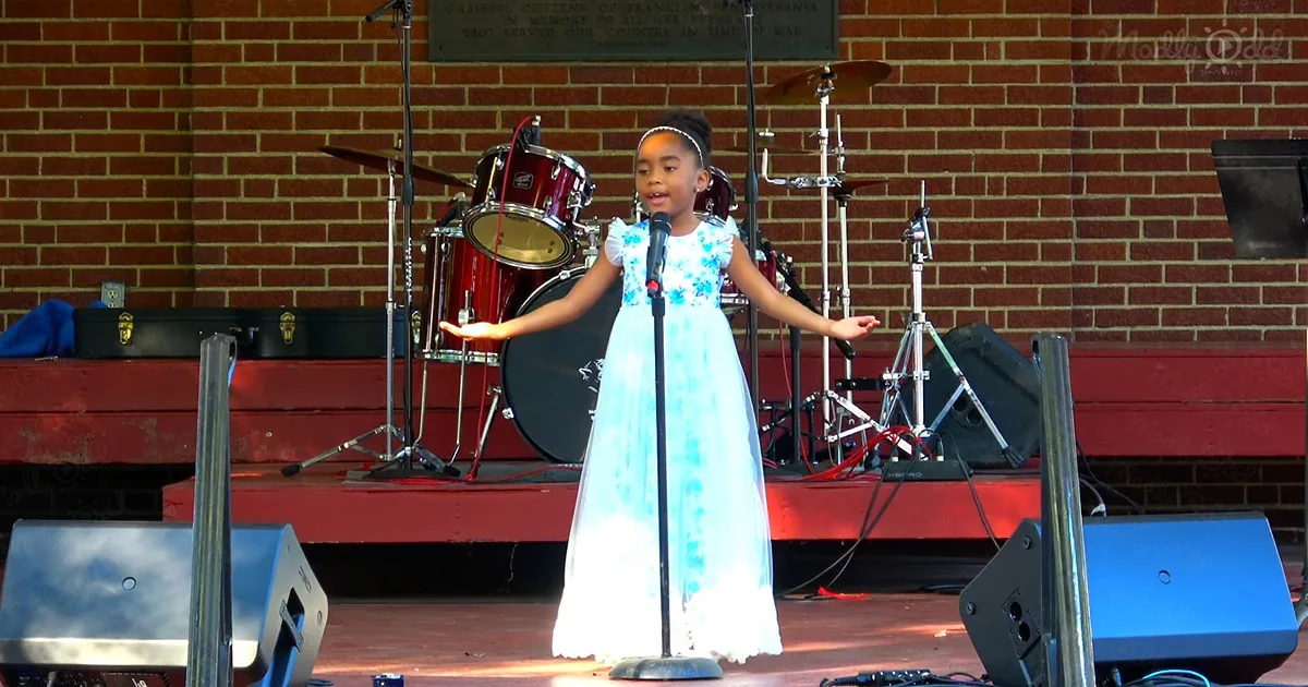 A young girl in a blue dress sings on stage with a microphone, surrounded by musical equipment.