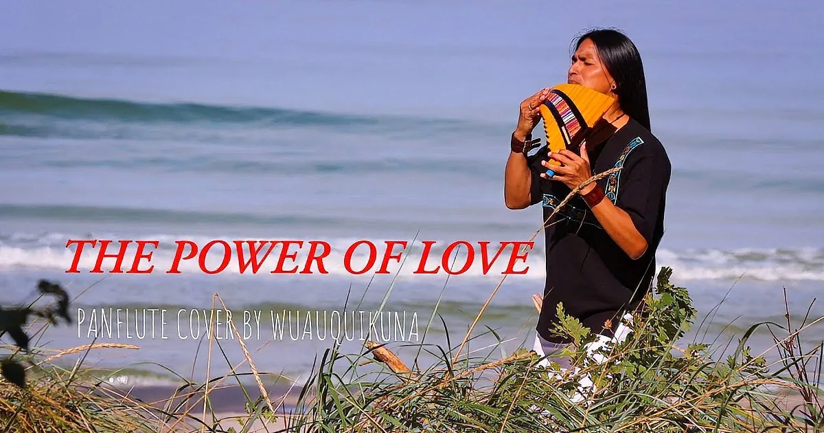 A person plays a colorful instrument near the ocean, with waves and grass in the background.