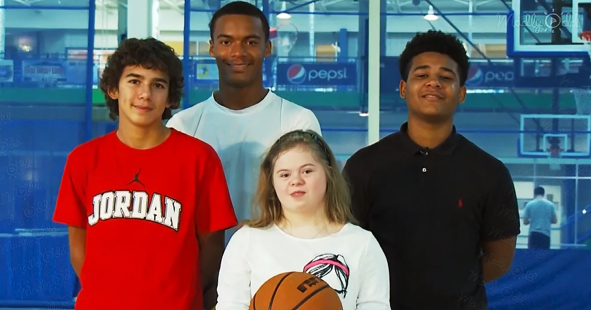 A girl with Down syndrome holds a basketball, flanked by three young men in a gym setting.