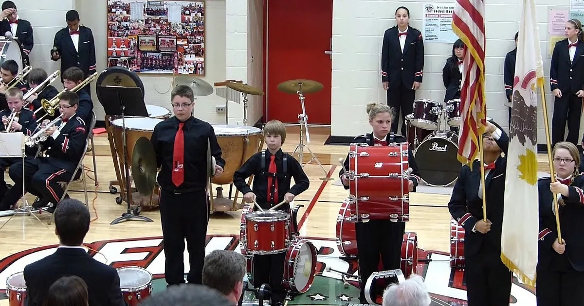 A young drummer in a black uniform with a red tie plays a red drum set in a school gymnasium.