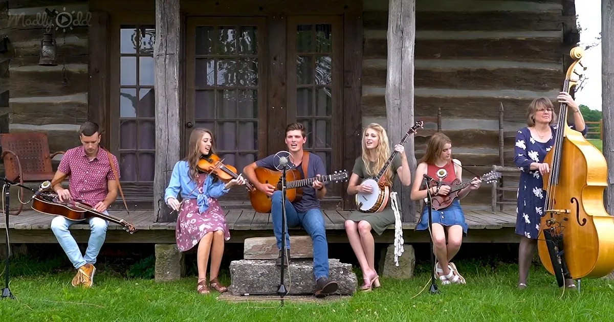 A group of musicians plays folk music on a porch in front of a rustic log cabin.