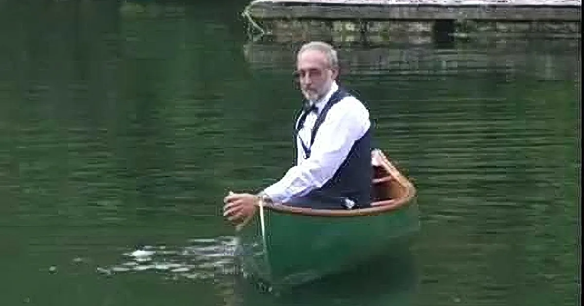 A man in a suit paddles a wooden canoe on calm water.