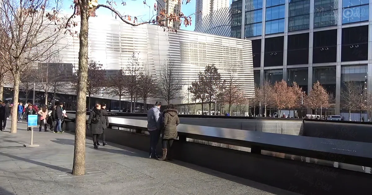 People walk near a reflective water feature in a cityscape with modern buildings and bare trees.