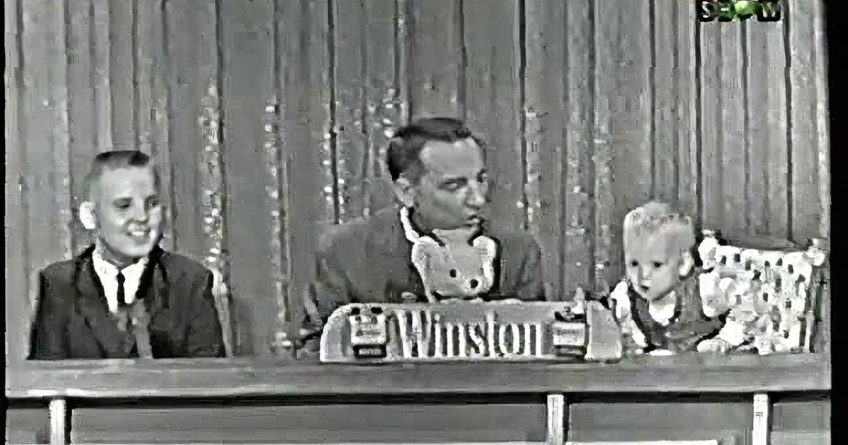 A man in a suit and tie is seated at a table, engaging with a young child who appears surprised.