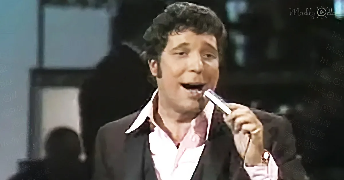 A man in a suit and pink shirt holds a microphone, singing with passion.