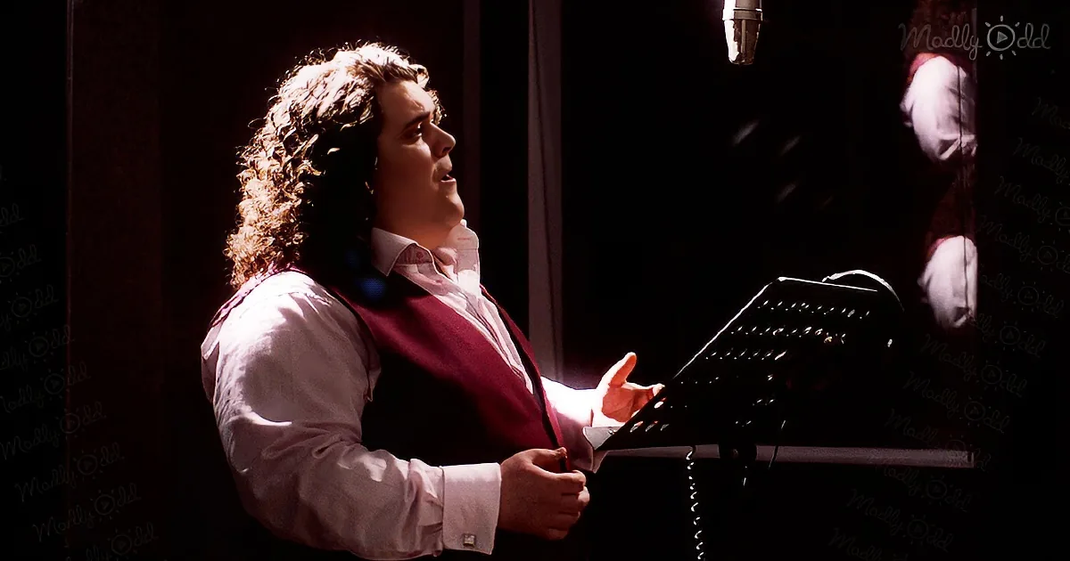 A man with curly hair, wearing a white shirt and red vest, sings into a microphone in a dimly lit room.