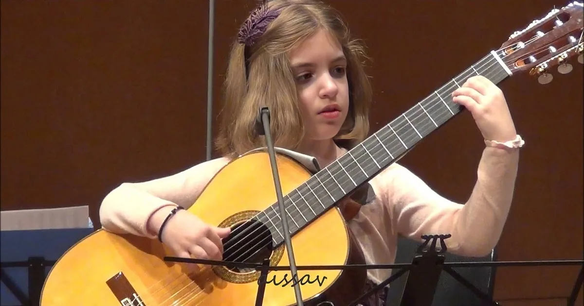 A young person plays a classical guitar in front of a microphone, with sheet music stands nearby.