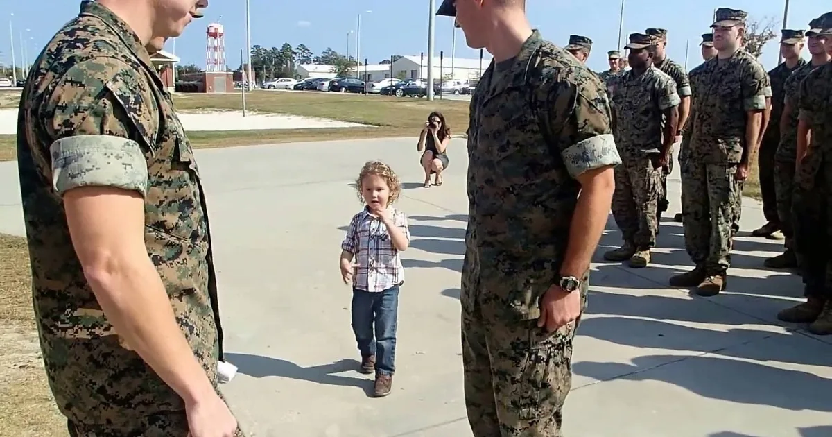 A young child in a plaid shirt helps a soldier in camouflage adjust his uniform.
