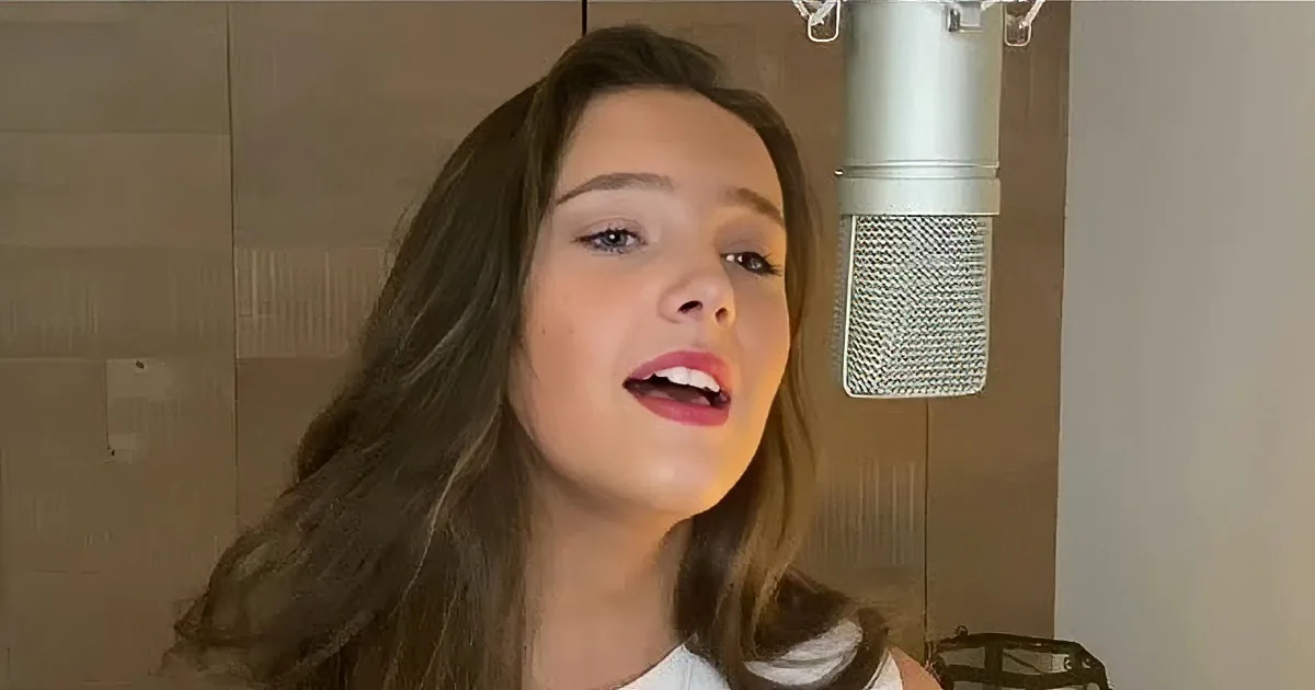 A woman sings into a microphone, wearing a white shirt, with a piano and warm lighting in the background.