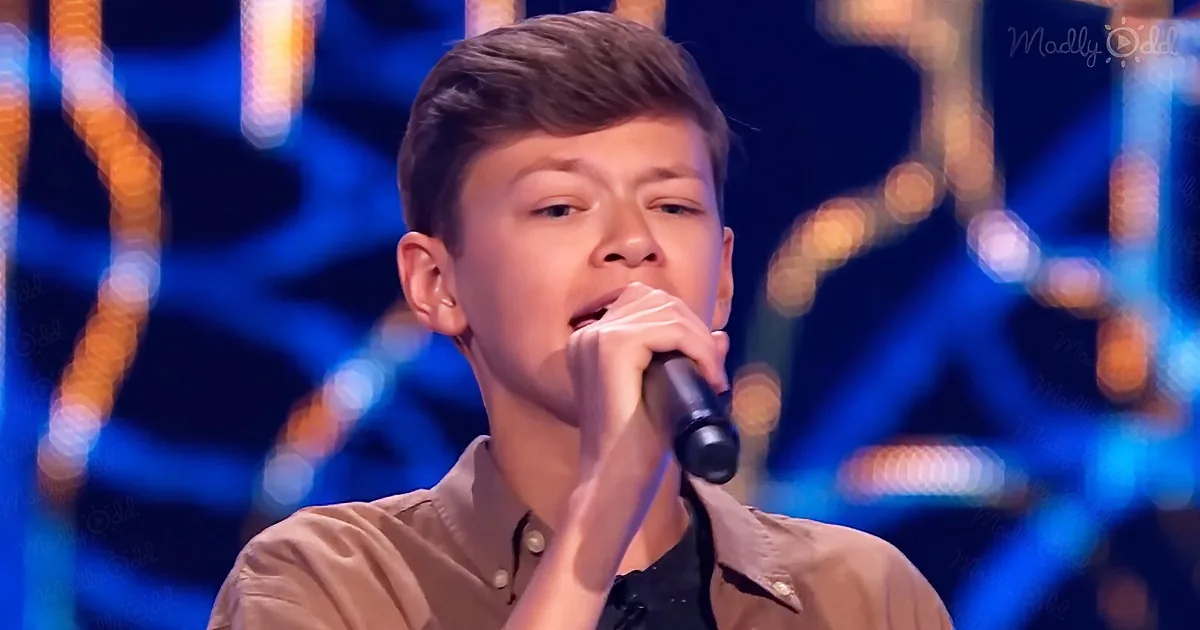 A young boy in a brown shirt sings into a microphone on a stage with blurred lights.