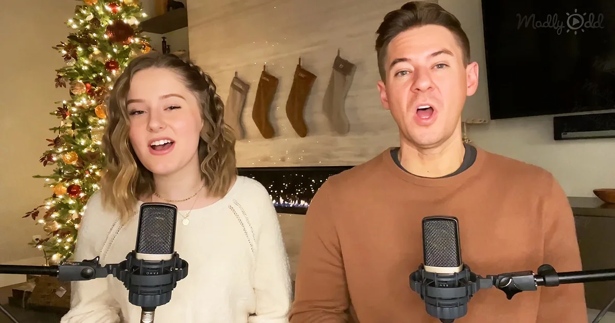 A man and woman sing into microphones in front of a Christmas tree and stockings.