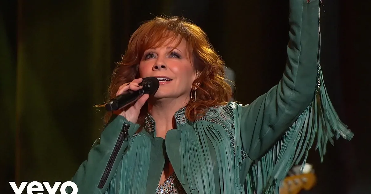 Reba McEntire sings passionately, holding a microphone, wearing a teal fringed jacket, with her hands raised.