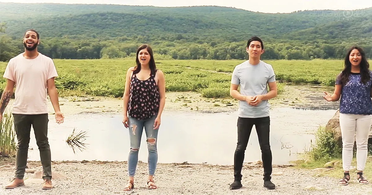 Four people stand by a river, smiling and singing, with a lush forest in the background.