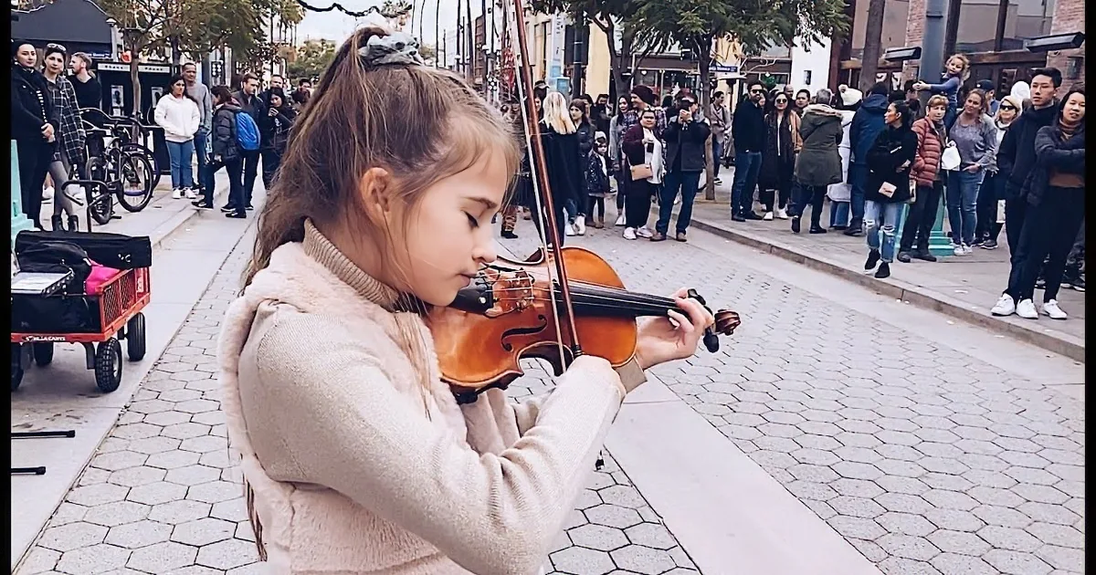 A young girl plays the violin on a city sidewalk, surrounded by onlookers.