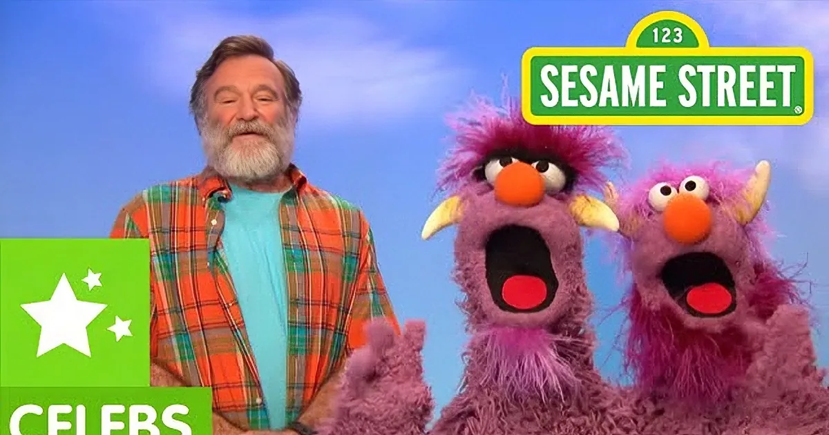 A man in a plaid shirt interacts with two pink, furry puppets against a blue sky backdrop.