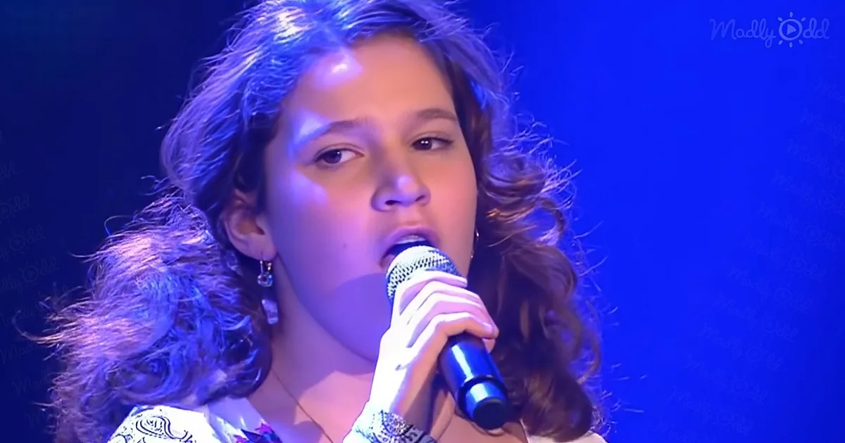 A person with curly hair sings into a microphone under blue stage lighting.