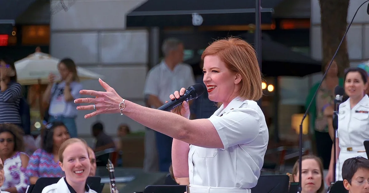 A woman in a white uniform sings into a microphone, raising her hand expressively.