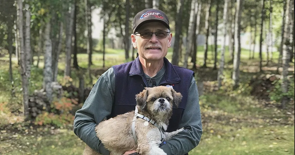 A man walks his small dog on a sidewalk, holding a leash and wearing a cap and sunglasses.