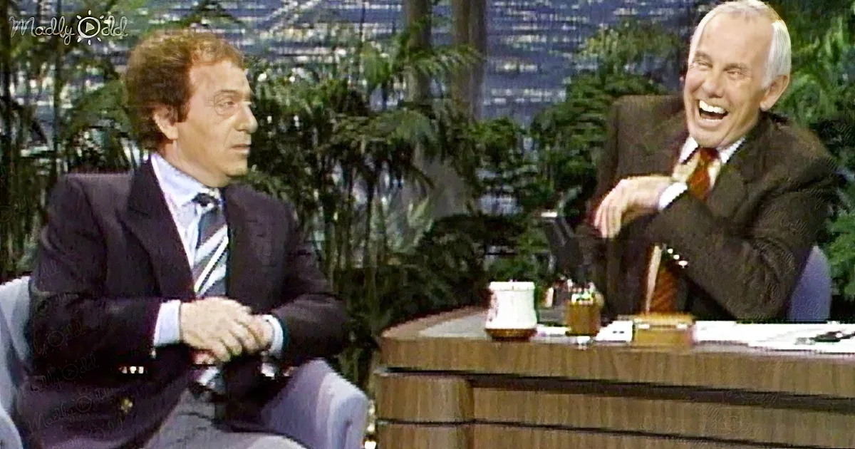 Two men in suits sit on a talk show set, one laughing, the other listening, with a desk and plants in the background.