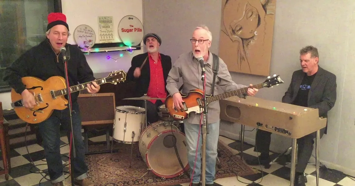 A band performs with a guitarist, drummer, bassist, and keyboardist in a cozy indoor setting.