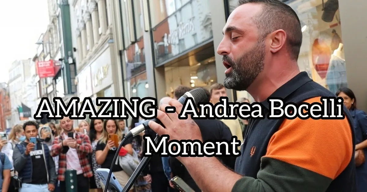 A man in a black and orange shirt sings into a microphone in front of a crowd.