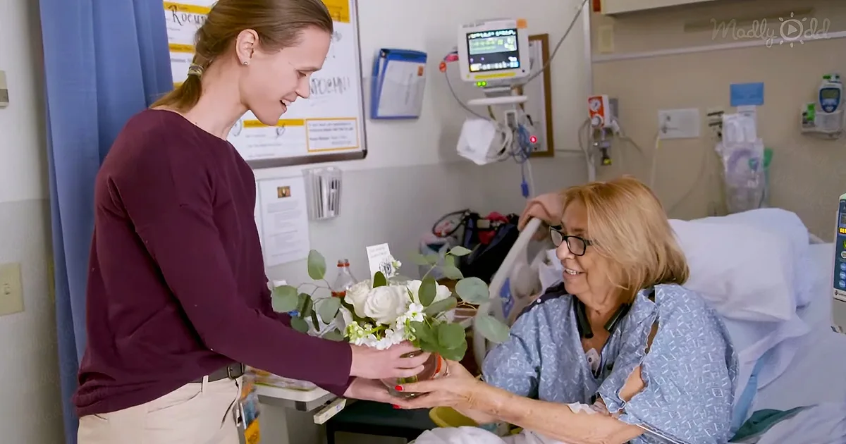 A woman in a hospital bed receives a bouquet of flowers from another woman.