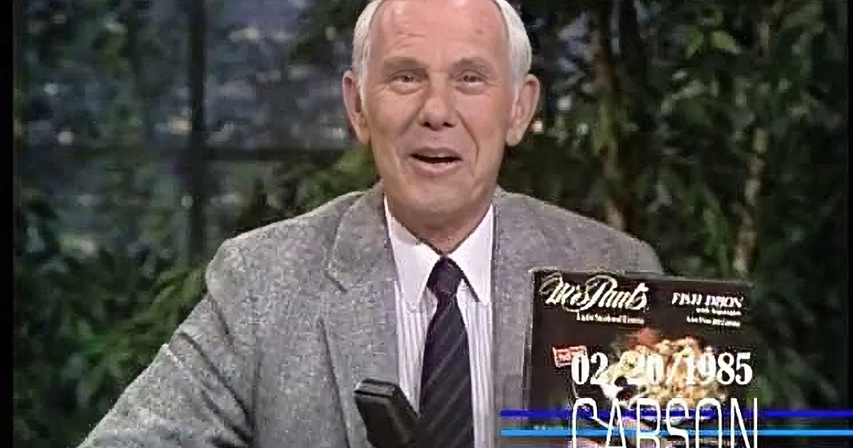An older man in a suit smiles while holding a microphone, with a backdrop of greenery and a cityscape.