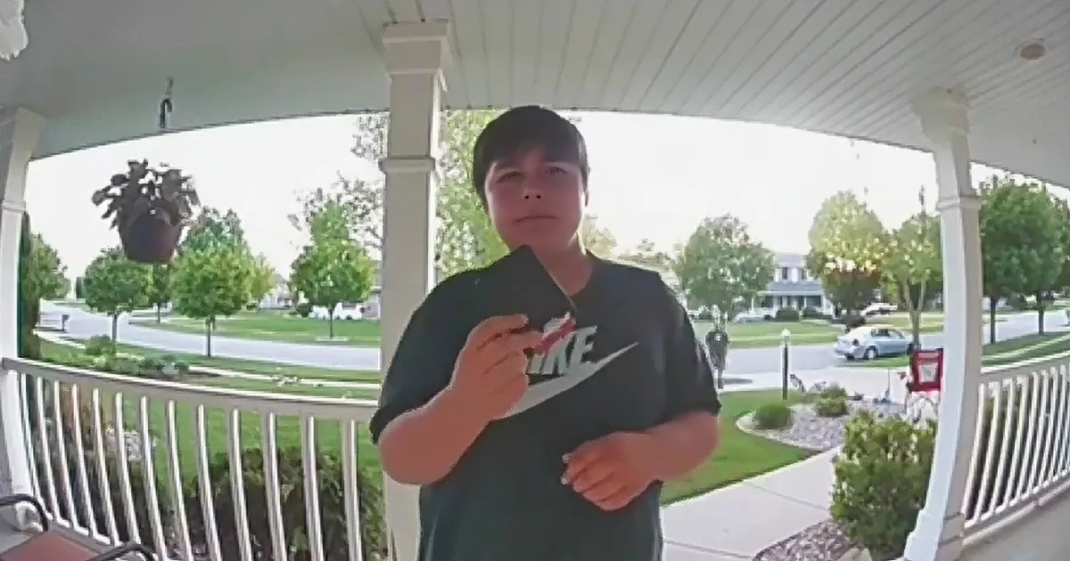 A person in a Nike shirt stands on a porch, holding a small object, with a suburban neighborhood in the background.
