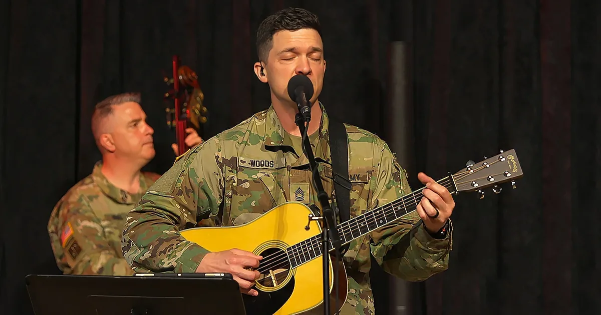 A soldier in camouflage plays guitar and sings into a microphone on stage.