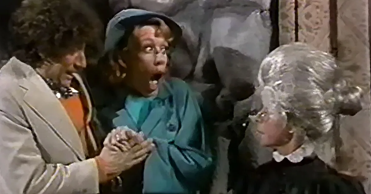 A woman in a teal coat and hat appears surprised, surrounded by others in a theatrical setting.