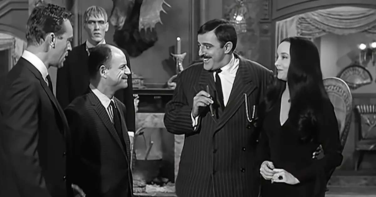 A man in a pinstripe suit holds a cigarette, smiling, with others in formal attire around him.