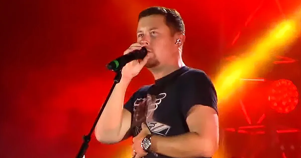 A man in a black t-shirt sings into a microphone on stage with red and yellow lights.