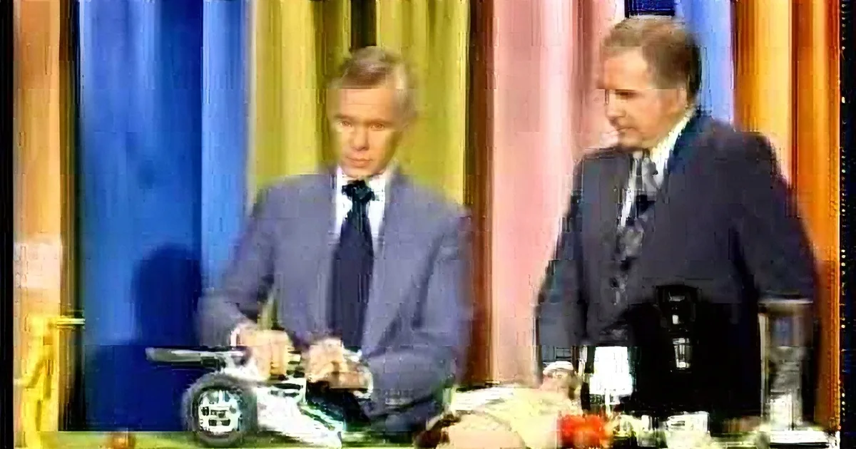 Two men in suits are signing documents at a table with microphones and a flag in the background.