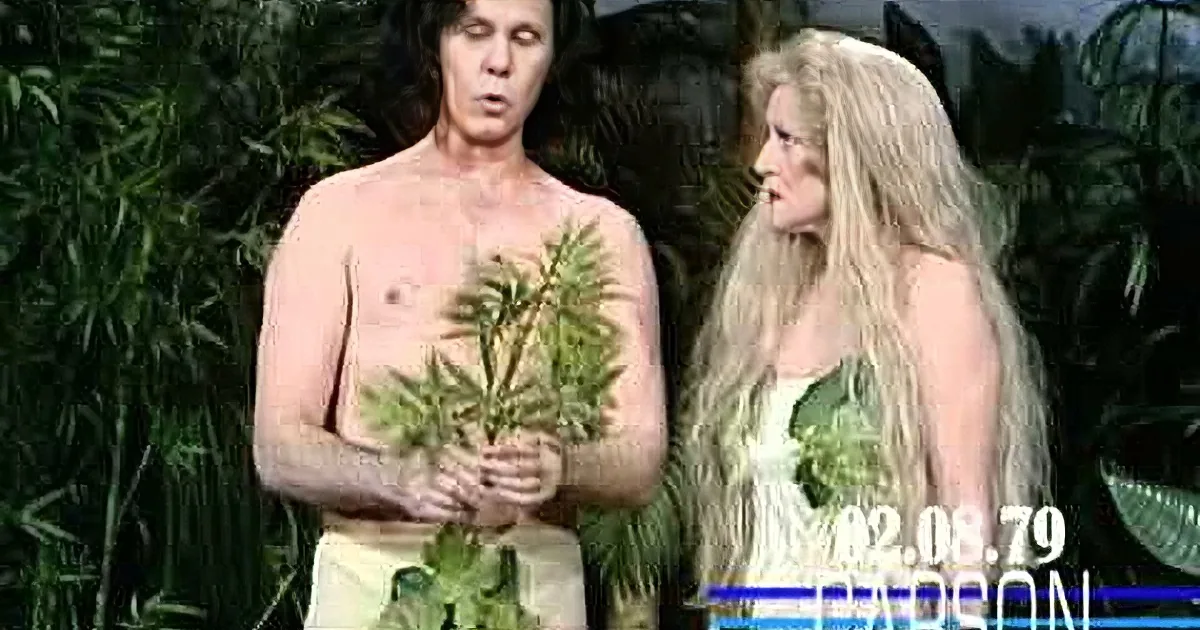 A man with long hair and no shirt stands in front of a woman with long blonde hair, both in a lush, green setting.