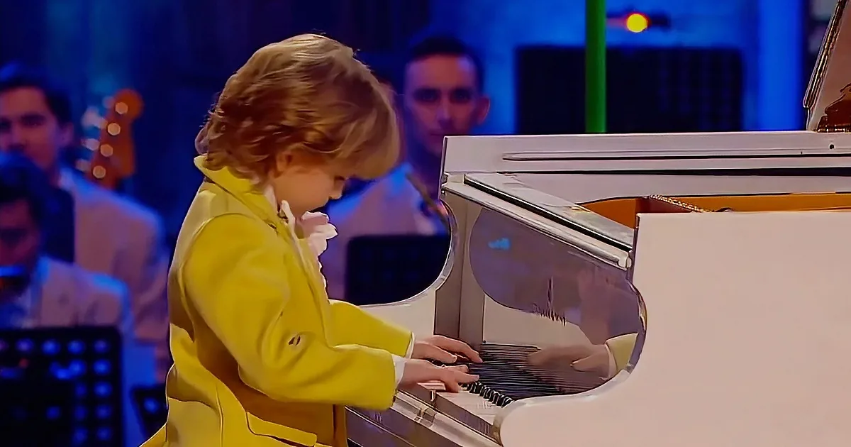 A young child in a yellow coat plays a white piano on a stage with musicians in the background.