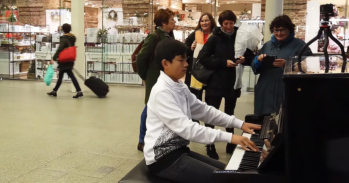 A young boy plays a piano in a shopping mall, surrounded by onlookers.