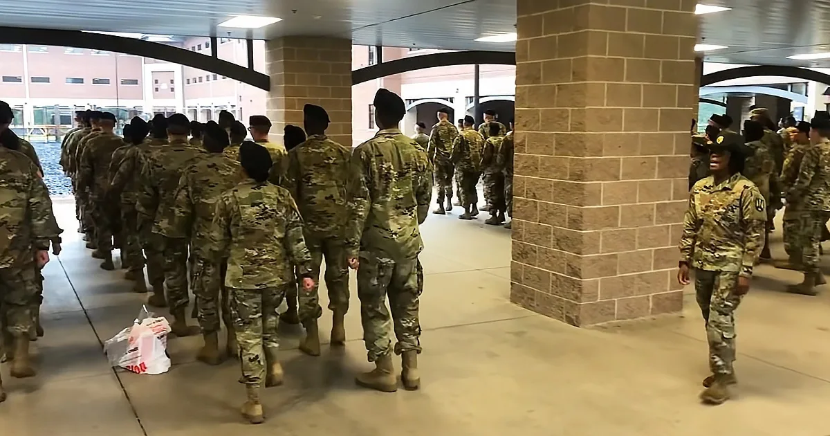 Soldiers in camouflage uniforms walk in formation through a building's hallway.