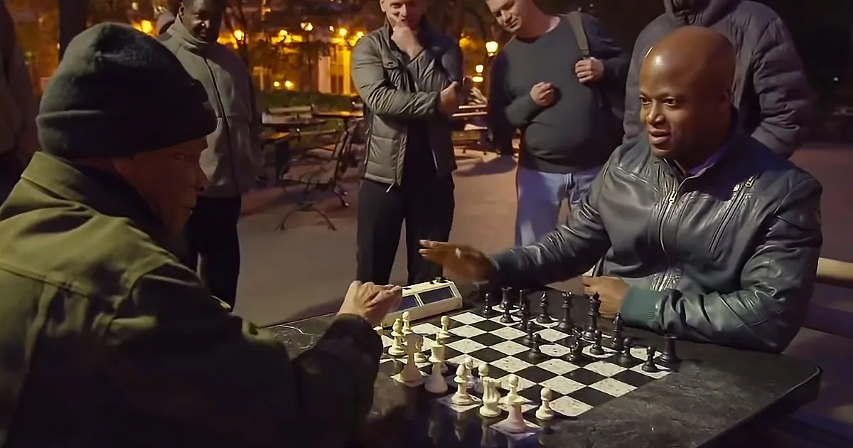 Two men play chess outdoors at night, surrounded by spectators.