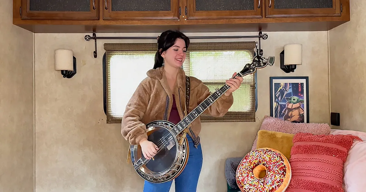 A person in a brown jacket plays a banjo in a cozy RV interior with a donut and Yoda poster.