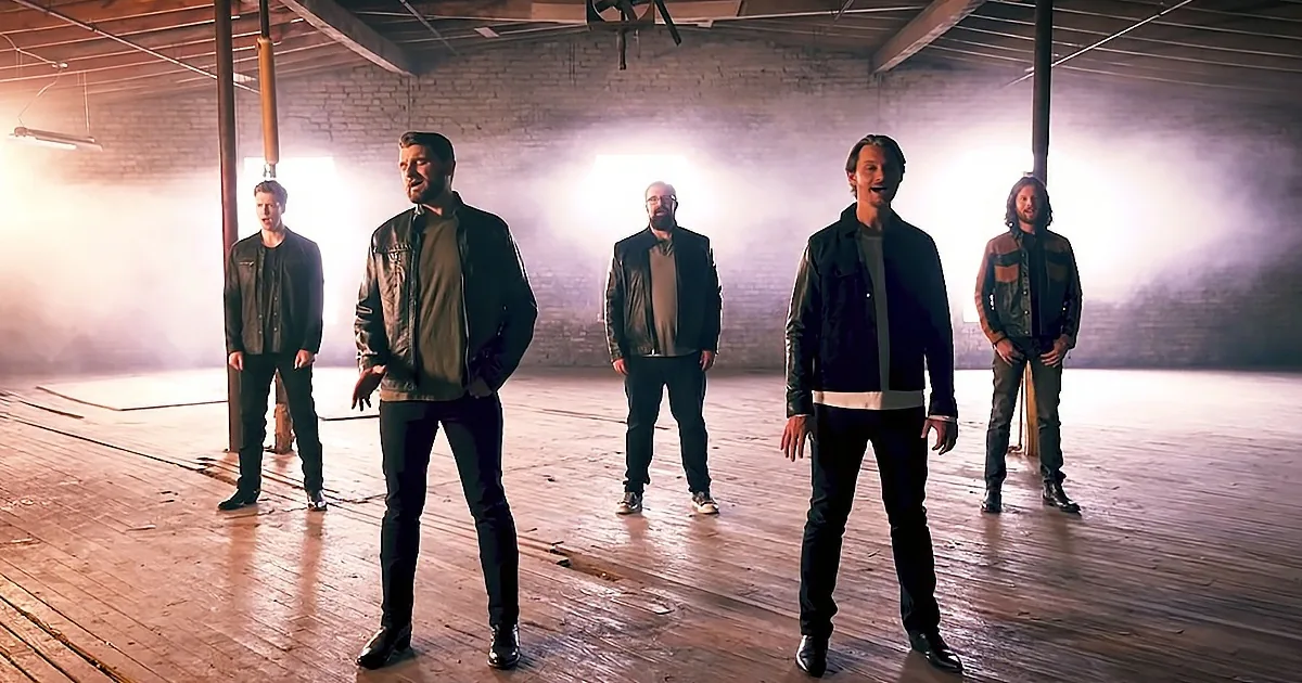 Five men in leather jackets stand in a smoky, dimly lit warehouse with wooden floors and brick walls.