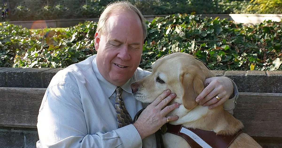 A man in a light shirt and tie pets a light brown Labrador Retriever on a bench.